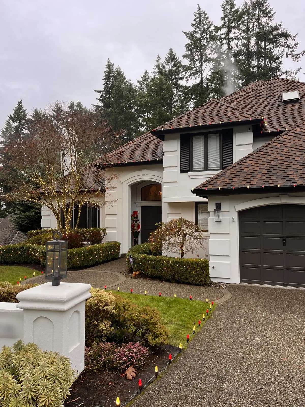 Warm and red holiday lights run along every roofline of a white stucco home, with a wrapped maple and lit pathway