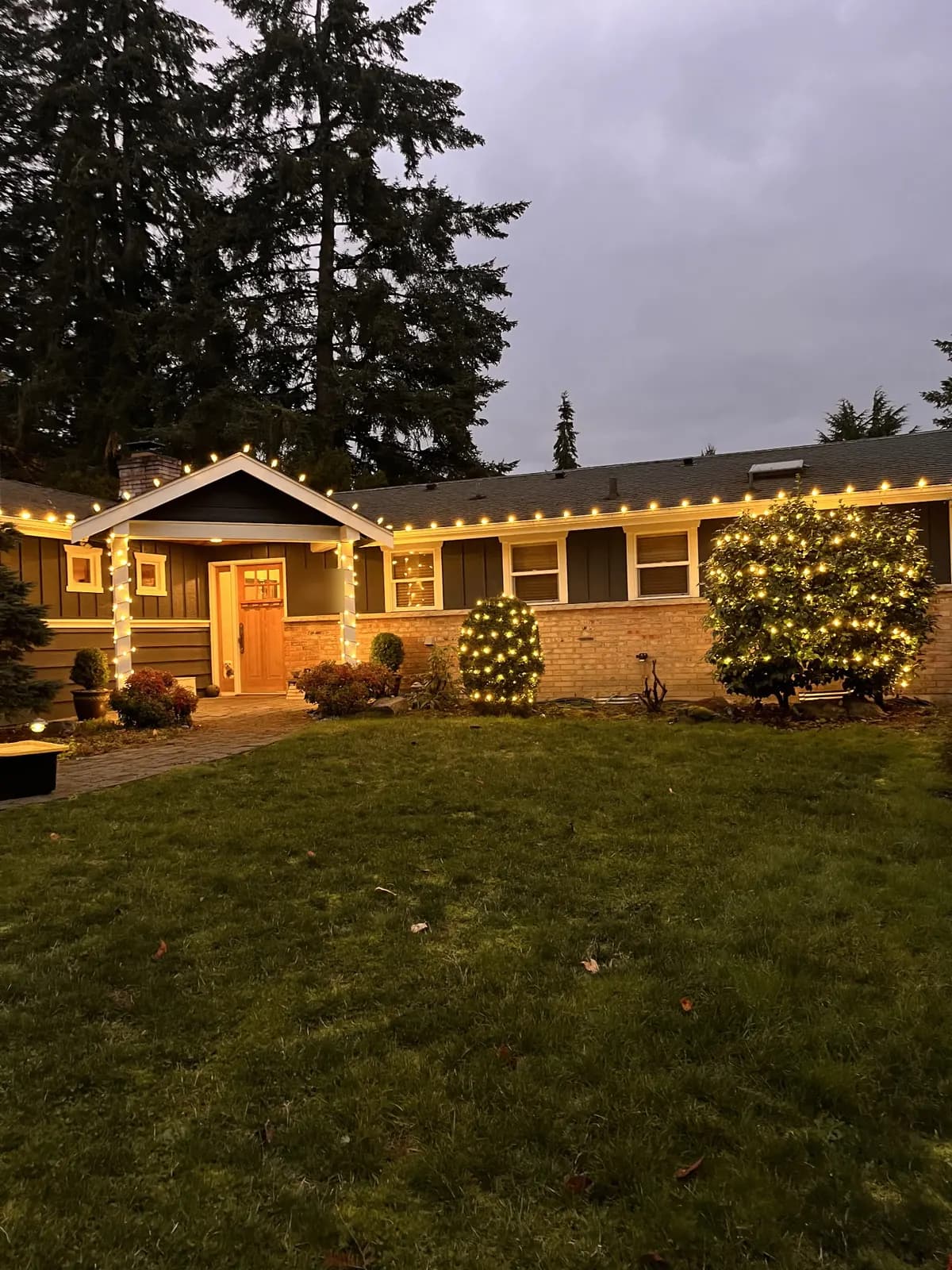 Warm holiday lights running the full eave line of a craftsman home at dusk, with lit shrubs below